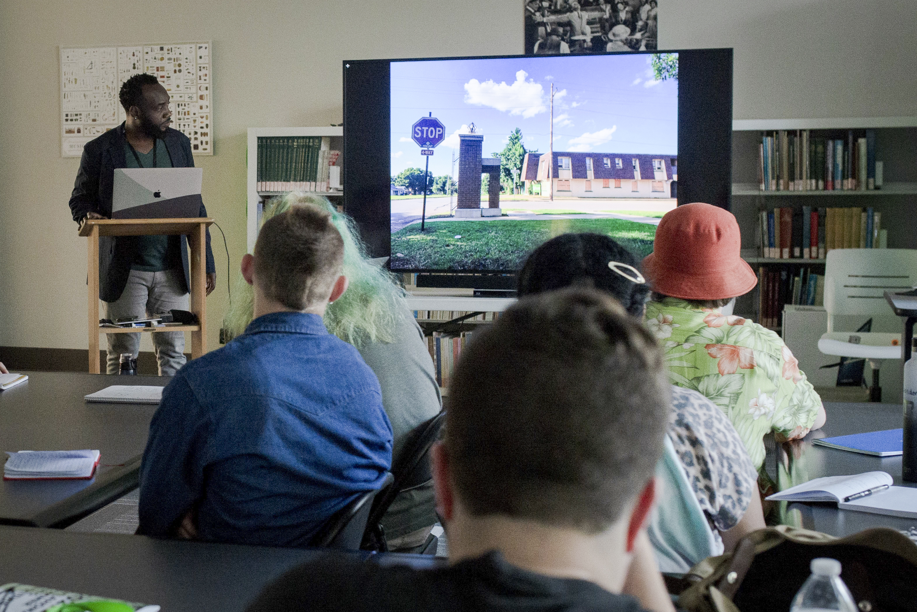 A man stands at a lectern in front of a group of students watching a film on a TV screen in a classroom setting.