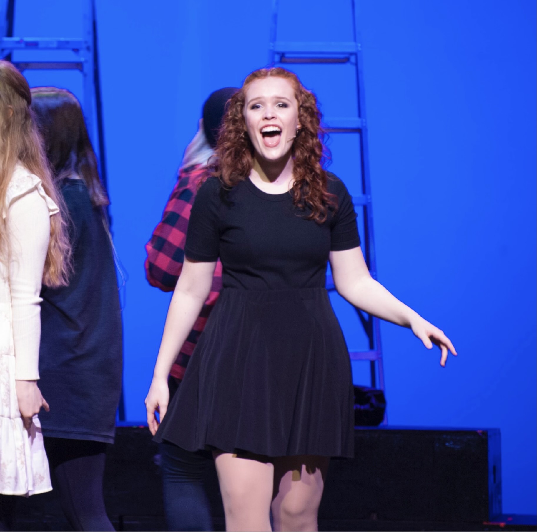A young person with long curly red hair, a black dress and a headset mic sings onstage in front of a blue backdrop, along with other performers whose faces are not in the frame