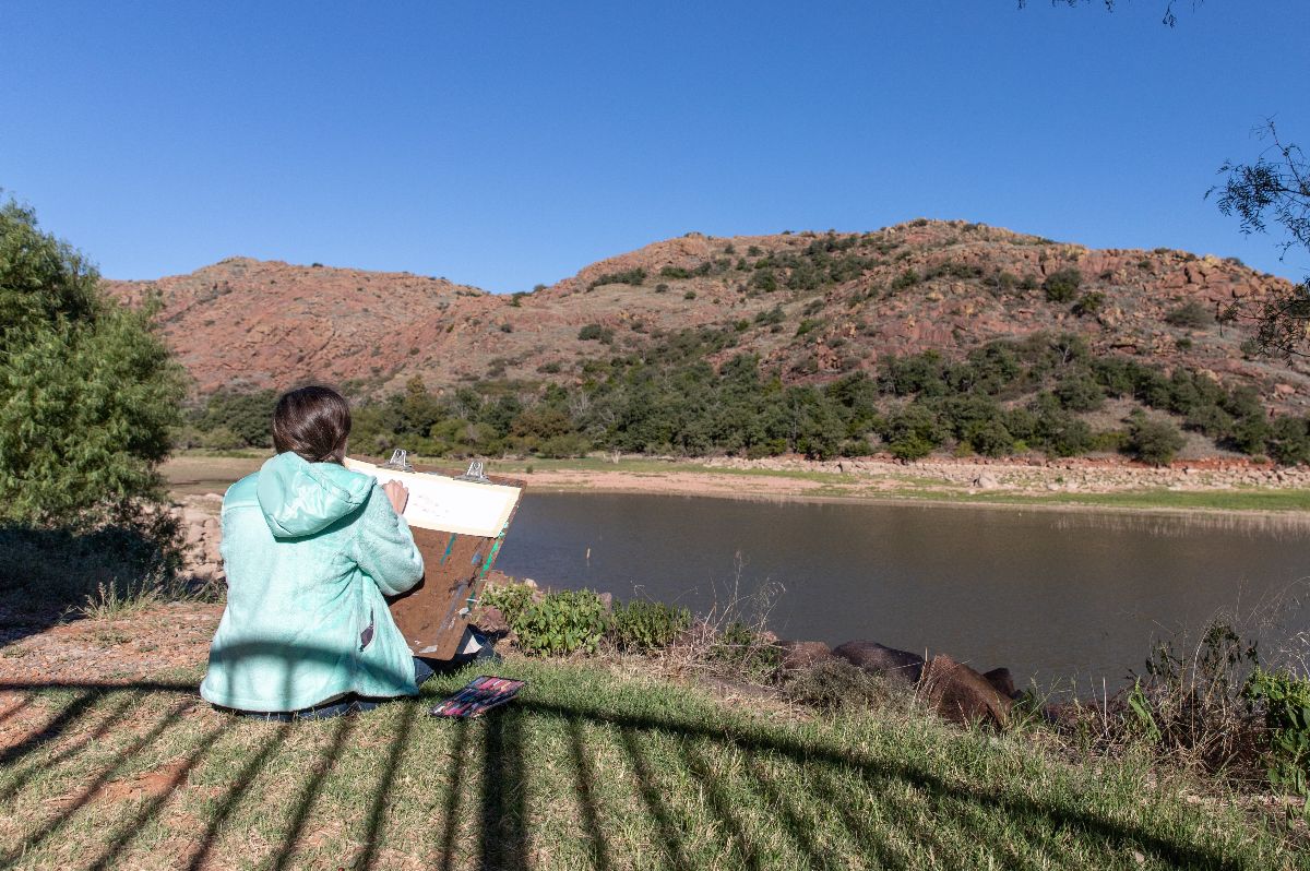 A person sits with their back to the camera, painting a scene of a lake and boulder-strewn mountains