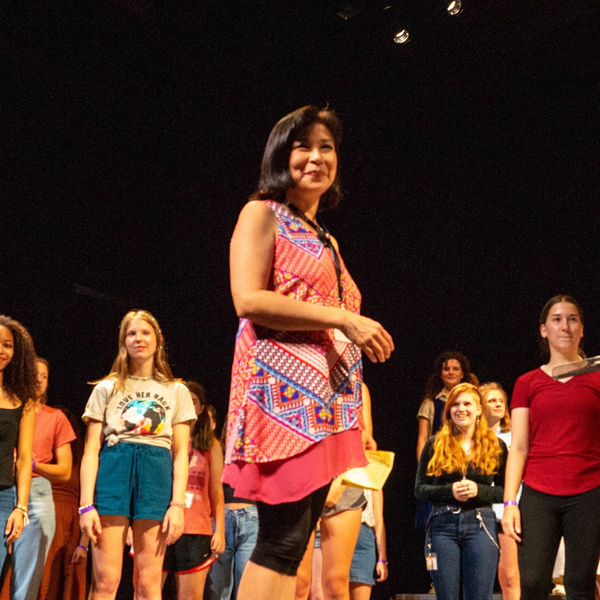 A person with dark hair and a headset mic stands in the spotlight on stage with rows of performers behind her