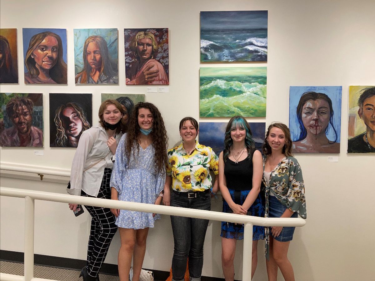 Five young people stand in front of paintings on canvas hung on a gallery wall