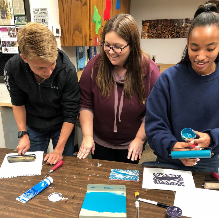 A teacher with glasses and long brown hair stands working with two students on a craft project