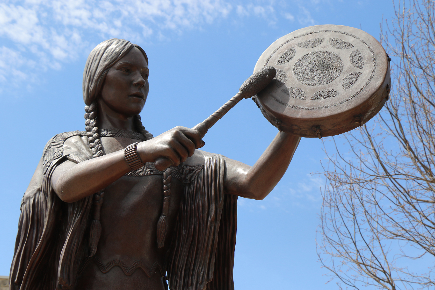 A statue depicting a woman in Native regalia as she strikes a hand drum with a mallet